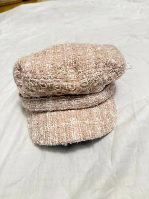 Beige/rosa tweedkeps från ALDO - Säljer en snygg beige/rosa keps i tweed från ALDO. Kepsen har en klassisk form med kort skärm, guldig knappdetalj på sidan och ett vävt tyg med inslag av vitt och guld. Perfekt accessoar för att lyfta din outfit.