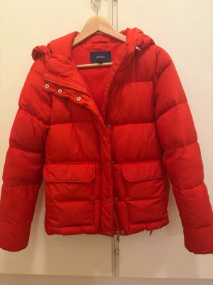 Röd pufferjacka från GANT XS - Snygg röd pufferjacka från GANT med huva och stora framfickor. Jackan har dragkedja och tryckknappar framtill samt quiltad design som ger en klassisk vinterlook. Perfekt för kalla dagar och riktigt varm.