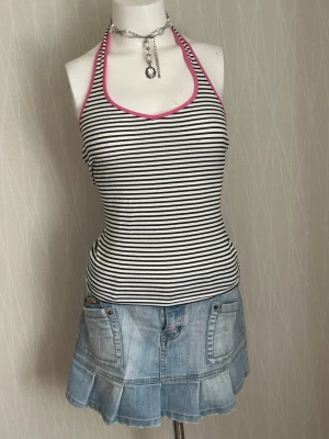Randig halterneck linnetopp - Snygg randig halterneck linnetopp med svarta och vita ränder och rosa kantband. Toppen är ärmlös och har en tight passform, perfekt för varma dagar. Materialet känns mjukt och stretchigt, vilket gör den bekväm att bära.