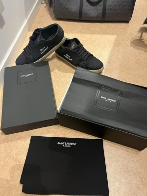 Svarta Saint Laurent sneakers - Svarta sneakers från Saint Laurent med vit broderad logga på sidan och guldfärgad text på plösen. Skorna har en klassisk låg siluett, svarta skosnören och beige sula. Tillverkade i canvasmaterial för en stilren och urban look.
