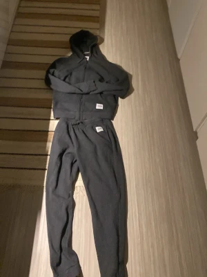 Mörk grå Andy sett - Mysigt svart loungewear-set från Herschel med matchande hoodie och byxor. Hoodien har dragkedja och fickor, byxorna har resår i midjan och muddar vid benslut. Perfekt för chill hemma eller när du vill vara bekväm.