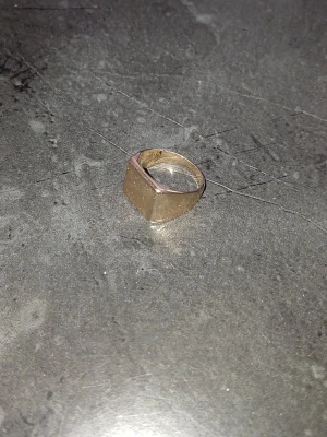 Klassisk klackring stämplad "14k"  från dödsbo  - Säljer en stilren signetring i guldfärgat metallmaterial med en fyrkantig, slät ovansida. Ringen har en enkel och tidlös design utan stenar eller dekorationer. Perfekt för dig som gillar minimalistiska smycken. Den är stämplad som 14k guld men jag tror inte att det är guld. Köp ej om du inte är införstådd med att det kanske inte är guld.