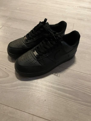Svarta Nike Air Force 1 sneakers - Klassiska helsvarta Nike Air Force 1 sneakers i läder med perforerad tå, svart sula och svarta skosnören. Swoosh-logga på sidan och metallbricka vid snörningen. Perfekta för en clean och stilren look. Typ helt nya använda typ 3-4 gånger pris kan diskutera alltid 