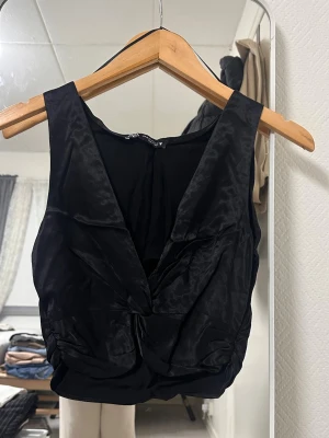 Svart omlott topp från Zara - Snygg svart omlott topp från Zara i glansigt satintyg. Toppen är ärmlös med djup v-ringning och twistad detalj framtill. Perfekt för att lyfta din outfit med en trendig touch.