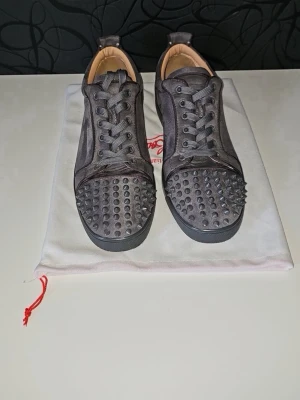 Christian Louboutin grå spikes sneakers - Snygga gråa sneakers från Christian Louboutin i mocka med ikoniska nitar på tån. Skorna har klassiska röda sulor och gråa skosnören. Perfekt för dig som vill sticka ut med en lyxig stil.