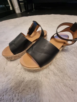 Svarta platåsandaler med rem - Snygga svarta sandaler med bred rem över foten och justerbar ankelrem. Sandalerna har en platåsula i flätad jute-look.