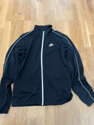 Svart träningsjacka från Nike - Svart träningsjacka från Nike med vit dragkedja och vita detaljer längs ärmarna. Jackan har hög krage, lång ärm och klassisk Nike-logga på bröstet. Perfekt för träning eller sportiga outfits.