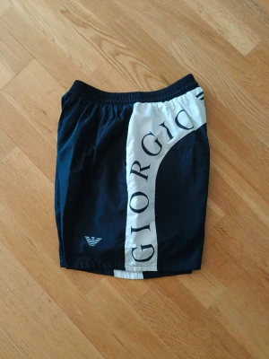 Giorgio Armani - Snygga marinblå shorts från Giorgio Armani med bred vit panel och stor logotyptext längs sidorna. Shortsen har elastisk midja och är perfekta för en avslappnad och trendig stil. Materialet känns lätt och sportigt.
