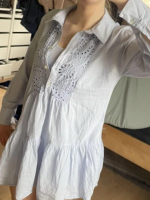 Ljusblå klänning från Zara - Ljusblå kort klänning från Zara med lång ärm och krage. Klänningen har broderade detaljer framtill, knappar vid bröstet och en luftig, volangig nederdel. Har en underklänning under som syns lite vid bysten på bild 1. Klänningen är i väldigt bra skick då den endast används fåtal gånger 