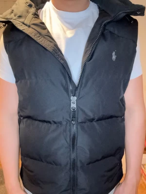 Svart dunväst med huva från Polo Ralph Lauren - Säljer en svart dunväst från Polo Ralph Lauren med huva och dragkedja framtill. Västen har ett klassiskt broderat logotyp på bröstet och är quiltad för extra värme. Perfekt för lager-på-lager och snygg till jeans eller joggers. Den passar alla olika stilar och sitter perfekt med en T-shirt eller en hoodie under när de är kalla kvällar. Den är använd ett få tall gånger. Denna modellen är svår och få tag på och är väldigt omtyckt. Om du ska köpa den ny så e du tvungen att betala en stor summa