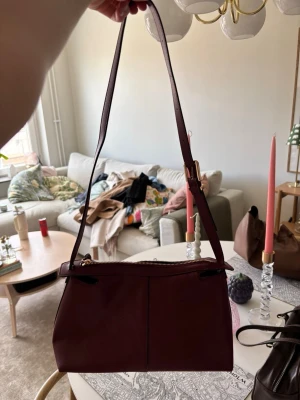 Vinröd axelväska i läder - Snygg vinröd axelväska med stilren design och justerbar axelrem. Väskan är tillverkad i slätt läder och har en praktisk dragkedja upptill. Perfekt för dig som vill ha en trendig och enkel väska som rymmer det viktigaste.