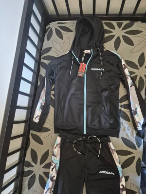 Askari tracksuit "SELASSIE" - Helt Ny tracksuit från Askari med luva och dragkedja. Setet har blå och vita camo-detaljer längs armarna och benen, samt logga på både jacka och byxa. Materialet är mjukt och stretchigt, perfekt för sport eller chill. Dragsko i midjan på byxorna för bästa passform. Jag köpte den från deras webbplats för 1700kr, och har aldrig haft på mig den.