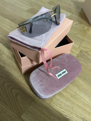 Miu Miu solglasögon med guldiga detaljer - Snygga solglasögon från Miu Miu med stora, rektangulära gråtonade glas och tunna guldiga bågar. Miu Miu-logga i guld på sidan. Kommer med rosa sammetsfodral och ask. Perfekt accessoar för att lyfta din stil.