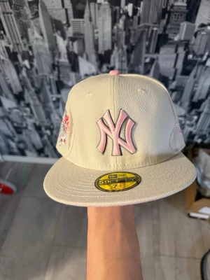 Beige New Era Yankees keps 59Fifty - Snygg beige New Era 59Fifty keps med rosa New York Yankees-logga framtill och broderade detaljer på sidan. Klassisk flat brim och MLB-logga bak. Tillverkad i bomullstyg och perfekt för dig som vill ha en clean och trendig look. 