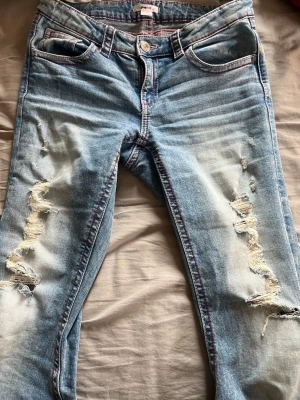 Ljusblå slitna jeans med brodyr - Ljusblå jeans från Gina tricot med slitningar framtill och broderade detaljer i rosa på bakfickorna. Jeansen har klassisk femficksmodell, knappgylf och kontrastsömmar. Perfekta för en avslappnad och trendig look. Inga defekter förutom de som visas på sista bilden vilket absolut inte är stort.