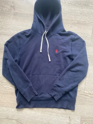 Mörkblå hoodie från Polo Ralph Lauren - Mörkblå hoodie från Polo Ralph Lauren med vit dragsko och röd broderad logga på bröstet. Klassisk känguruficka framtill och mjukt material som känns skönt mot huden. Perfekt för en avslappnad och stilren look.