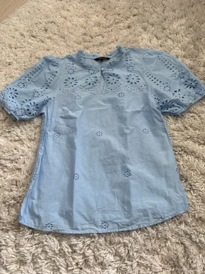 Ljusblå blus med broderade detaljer - Söt ljusblå blus med korta ärmar och broderade blommönster. Blusen har en liten volang vid halsen och en knappöppning framtill. Perfekt för en fräsch och somrig look.