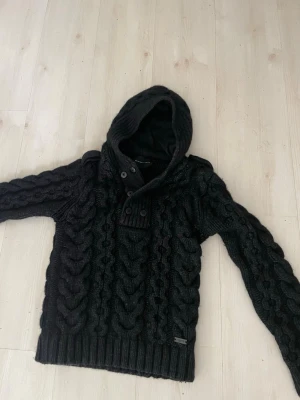 Svart kabelstickad hoodie Selected Homme - Mörkgrå kabelstickad hoodie från Selected Homme med stora flätmönster, knappar vid halsen och ribbade muddar. Perfekt för kalla dagar med sin mysiga huva och snygga detaljer.
