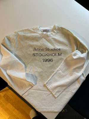 Vit långärmad Acne Studios - Vit långärmad  från Acne Studios med tryck 'Acne Studios STOCKHOLM 1996' på bröstet. Klassisk rund halsringning och avslappnad passform. Perfekt för en clean och stilren look.