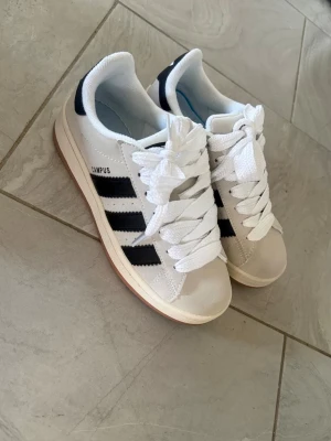 Adidas Campus vita sneakers - Fräscha Adidas Campus sneakers i vitt med svarta ränder och gummisula. Klassisk låg modell med breda vita skosnören och Campus-tryck på sidan. Perfekta för en clean och sportig stil. Kommer med originalkartong. Jag har använt de en gång.