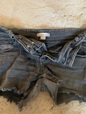 Lågmidjade Grå jeansshorts 🩶 - Säljer ett par grå jeansshorts från Gina med rå, fransig kant och klassisk femficksmodell. Shortsen har dragkedja och knapp framtill samt en avslappnad passform. Perfekta för varma dagar och en cool streetstil.