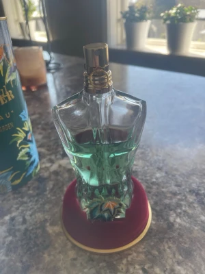 Jean Paul Gaultier Le Beau Paradise Garden - Jean Paul Gaultier Le Beau Paradise Garden är en parfym i en unik flaska formad som en manlig torso. Flaskan är genomskinlig med ljusgrön vätska och har detaljer i guld. Kommer i en dekorativ grön tub med tropiska motiv och guldtext.