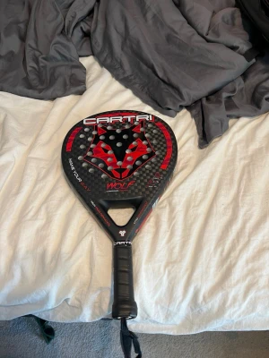 Padelracket Cartri Wolf svart/röd - Säljer ett padelracket från Cartri, modell Wolf. Racketen är svart med röda detaljer och har en cool vargdesign på träffytan. Tillverkat i kolfiber med perforerad yta och greppvänligt handtag. Perfekt för dig som gillar padel och vill sticka ut på banan.