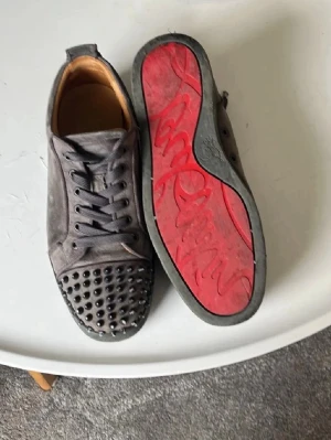 Gråa Louboutin sneakers med nitar - Säljer ett par gråa sneakers från Christian Louboutin med ikoniska röda sulor och coola svarta nitar på tån. Skorna har snörning och är tillverkade i mocka för en lyxig känsla. Perfekta för dig som vill sticka ut med en edgy och exklusiv stil.