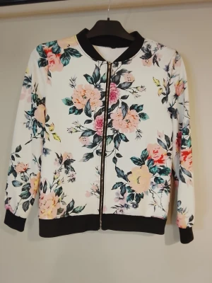 Vit blommig ziptröja med svarta detaljer NY - Snygg ziptröja med vit bas och färgglatt blommönster i rosa, gul och grönt. Tröjan har svarta muddar och krage samt dragkedja framtill. Perfekt för dig som gillar att sticka ut med mönster och färg. Riktigt snygg & fräsch vår/sommar kofta i nyskick.