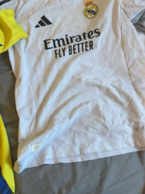 Real Madrid matchtröja Adidas vit - Säljer en vit Real Madrid matchtröja från Adidas med klubbmärke och sponsortryck på bröstet. Tröjan har svarta detaljer och är tillverkad i ett lätt och ventilerande material, perfekt för fotbollsträning eller match.
