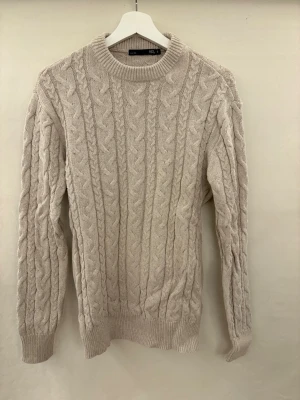 Beige kabelstickad tröja  - Kabelstickad tröja i beige från lager 157 med rund halsringning och ribbade muddar vid ärmslut och nederkant. Tröjan har ett klassiskt flätmönster och är perfekt för lager på lager under kyliga dagar. Har inte använt sedan den är köpt på grund av fel storlek. Pris kan diskuteras!