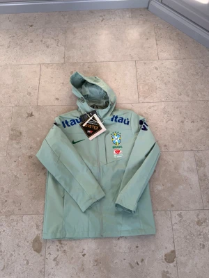 Nike Brasil vindjacka med huva - Ljusgrön vindjacka från Nike med huva och dragkedja. Jackan har Brasilien CBF-logga, sponsortryck och är tillverkad i GORE-TEX-material. Perfekt för dig som gillar sportig stil och vill visa stöd för Brasilien.