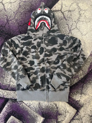 Grå camo hoodie med hajmotiv - Säljer en grå camo hoodie från BAPE med hel dragkedja och ikoniskt hajmotiv på huvan. Hoodien har ribbade muddar och fickor framtill. Perfekt för dig som gillar streetwear och vill sticka ut med en unik design.