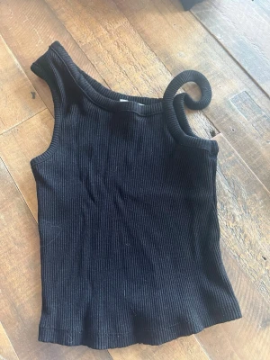 Svart topp från NA-KD - Snygg svart ribbad topp från NA-KD i oneshoulder-modell. Toppen är ärmlös och har en tight passform med stretchigt material, perfekt för att skapa en trendig look. Passar dig som gillar stilrena och moderna plagg.