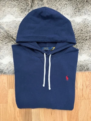 Marinblå Polo Ralph Lauren Hoodie - Storlek L - Hej! Säljer en klassisk marinblå Polo Ralph Lauren hoodie i storlek L med röd logga. 🐎 Tröjan är i använt skick med en del nopprighet, priset är därefter! ✨ 100% äkta – QR-koden finns, men ger meddelandet "authentication check". Detta är en säkerhetsspärr från Ralph Lauren som triggas när koden skannats från flera platser tidigare. Det är ett kvitto på att den varit populär på andrahandsmarknaden, inte att den är falsk. ✅ 