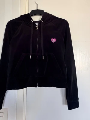 Svart velourhoodie med rosa hjärta - Svart hoodie i mjuk velour från Juicy Couture med dragkedja, huva. På bröstet sitter ett litet rosa hjärta i glittrande stenar. Perfekt för en chill och trendig look.