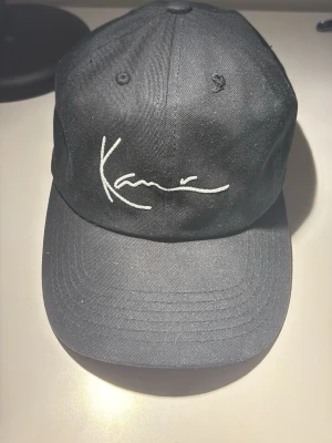 Svart keps från Karl Kani - Snygg svart keps från Karl Kani med vit broderad logga framtill. Klassisk böjd skärm och justerbart spänne baktill i metall. Tillverkad i bomullstyg som andas och har broderade ventilationshål för extra komfort.