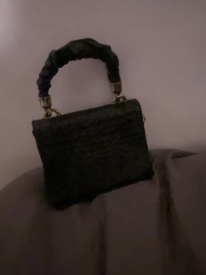 Svart handväska från Dolce & Gabbana - Snygg svart handväska i krokopräglat material från Dolce & Gabbana. Väskan har ett robust handtag klätt i tyg och stora dekorativa guldfärgade DG-bokstäver framtill. Perfekt accessoar för att lyfta din outfit.