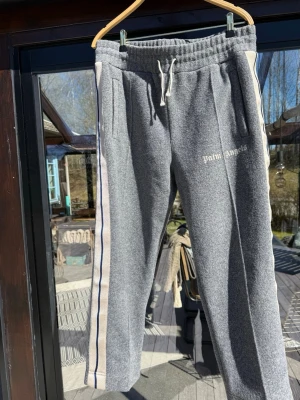 Grå Palm Angels byxor - Grå mjukisbyxor från Palm Angels med vita och blå ränder längs sidorna. Byxorna har resår i midjan med dragsko och två sidofickor. Broderad logga på vänster ben. Perfekta för en chill och avslappnad stil. Perfekt skick, inga defekter. Skriv vid frågor!