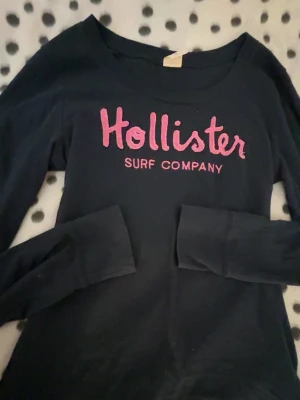 Svart långärmad tröja Hollister - Svart långärmad tröja från Hollister med rosa broderad text 'Hollister Surf Company' på bröstet. Tröjan har rund halsringning och är tillverkad i mjukt bomullsmaterial. Perfekt för en avslappnad och stilren look.