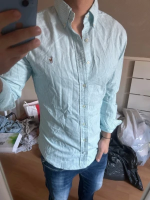 Randig skjorta från Ralph Lauren - Snygg ljusblå och vit randig skjorta från Ralph Lauren med klassisk button-down krage och broderad logga på bröstet. Skjortan har lång ärm och knäppning framtill. Perfekt till jeans för en clean och fräsch look.
