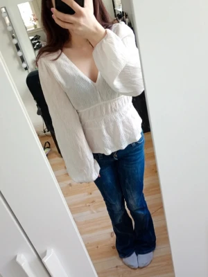 Vit v-ringad blus med ballongärmar - Säljer en stilren vit blus med djup v-ringning och ballongärmar. Blusen har en lätt rynkad struktur och markerad midja som ger en snygg siluett. Perfekt att matcha med jeans eller kjol för en trendig look ☺️