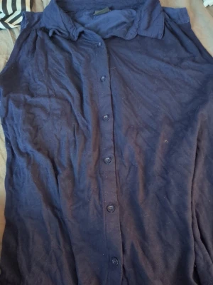 Mörkblå ärmlös blus med krage - Stilren mörkblå blus med knappar framtill och klassisk krage. Blusen är ärmlös och har en avslappnad passform, perfekt att matcha med jeans eller kjol. Materialet känns mjukt och lätt, vilket gör den skön att bära under varma dagar.
