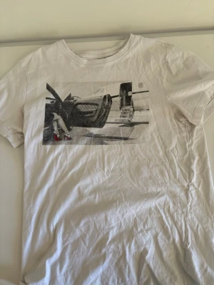 Vit t-shirt med tryck från Mister Tee - Vit t-shirt från Mister Tee med ett svartvitt tryck på bröstet som visar en bil och ett par ben i röda klackar. Klassisk rund hals och korta ärmar. Perfekt för dig som gillar streetstyle och coola motiv.