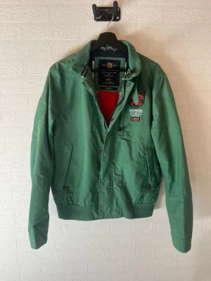 Grön vindjacka från Morris Heritage - Stilren grön jacka från Morris Heritage med broderad logga på bröstet och ärmen. Jackan har ribbade muddar, ståkrage och två snedställda fickor framtill. Storleken är medium. Skriv vid frågor, jag svarar snabbt!