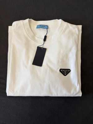 Vit t-shirt från zalando - Snygg vit t-shirt från zalando med klassisk rund hals och korta ärmar. På bröstet sitter zalando ikoniska svarta trekantslogga i metall. Tillverkad i mjuk bomull för en skön känsla. Perfekt för dig som gillar stilrena T-shirt.
