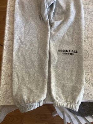 Grå Essentials Fear of God mjukisbyxor - Säljer ett par grå mjukisbyxor från Essentials Fear of God. Byxorna har resår i bensluten och midjan samt snörning för bästa passform. Tillverkade i mjukt material och har en stilren logga tryckt på benet. Perfekta för en avslappnad och trendig look.