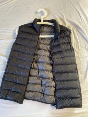 Svart dunväst från Uniqlo - Snygg svart dunväst från Uniqlo med lättviktigt material och quiltad design. Västen har hög krage och dragkedja framtill, perfekt för lager-på-lager-stil. Tillverkad i ett glansigt tyg som ger en modern look.