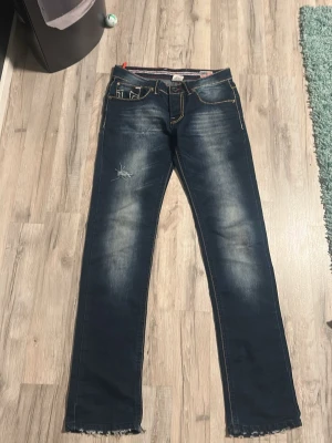 Mörkblå True Religion jeans - Säljer ett par mörkblå jeans från True Religion med slitningar på låret och fransade benslut. Jeansen har kontrastsömmar, klassiska fem fickor och stora broderade bakfickor med logga. Materialet är denim i bomull.