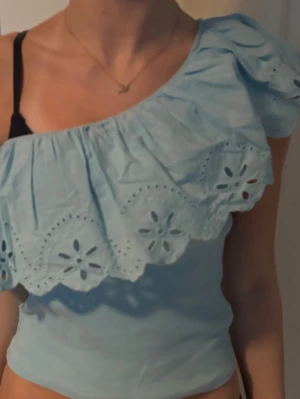 Ljusblå oneshoulder topp med volang - Söt ljusblå topp med oneshoulder-design och bred volang med broderade blommönster. Toppen är ärmlös på ena sidan och har en romantisk känsla, perfekt för varma dagar.